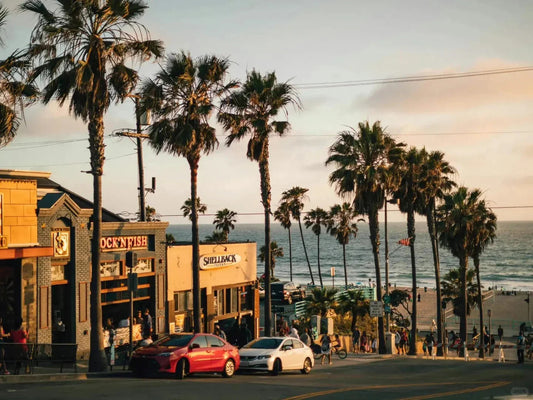 Beach street scene with palm trees, shops, parked cars, and people at sunset by the ocean.